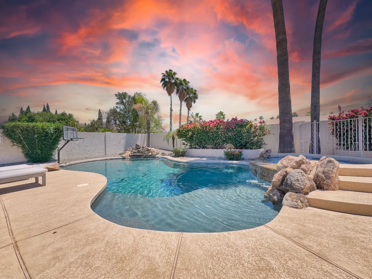 The image showcases a sparkling swimming pool surrounded by lush palm trees and manicured landscaping, set against a vibrant Arizona sunset sky. The inviting backyard features areas for relaxing, outdoor dining, and a basketball hoop for fun and games, creating an atmosphere perfect for memorable gatherings.