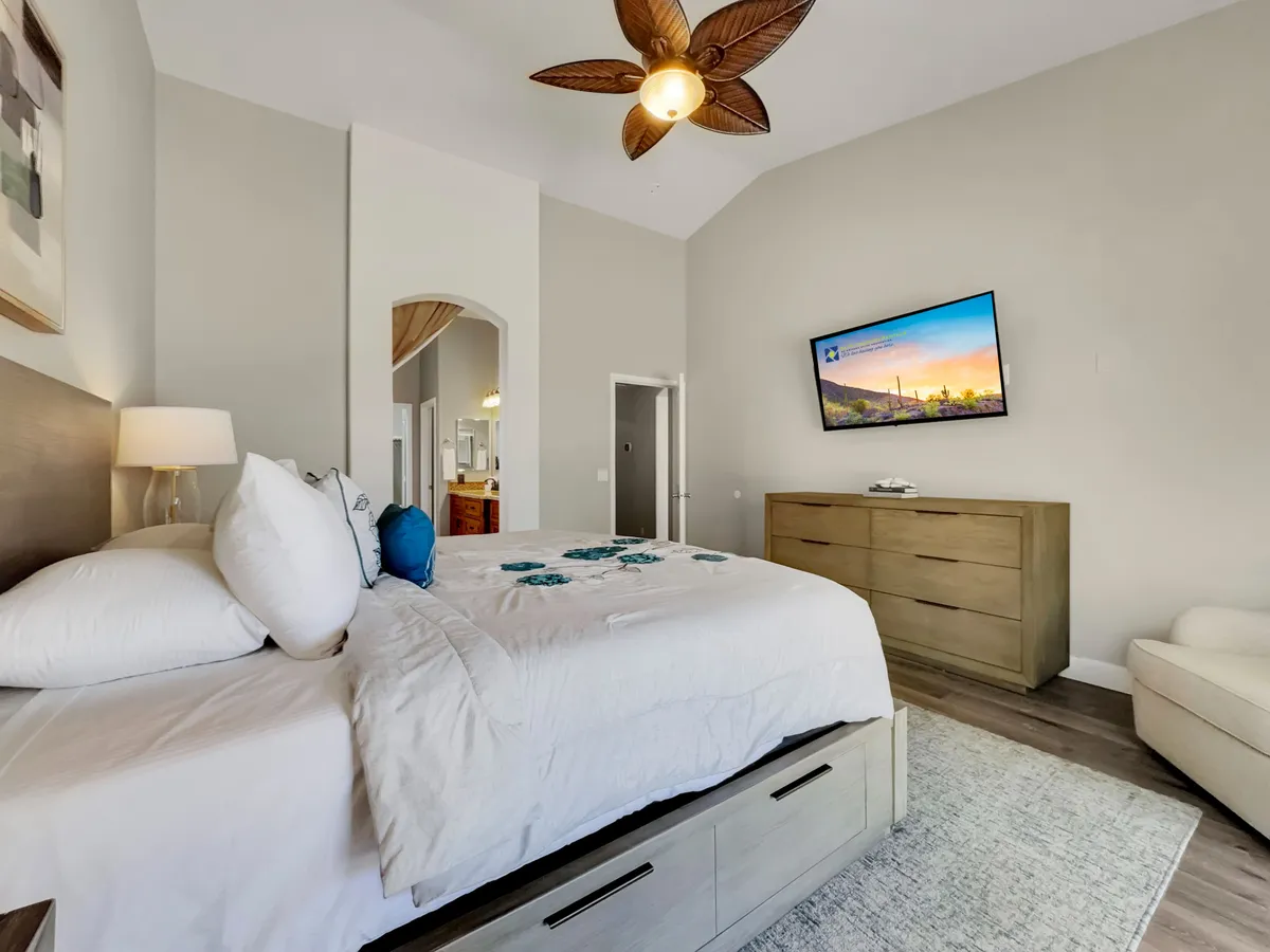 A queen bedroom featuring a bed with decorative pillows, calm artwork, a ceiling fan, and a flat-screen TV. The room is softly lit, with a dresser and soothing décor that create a tranquil, inviting space for relaxation and comfort.