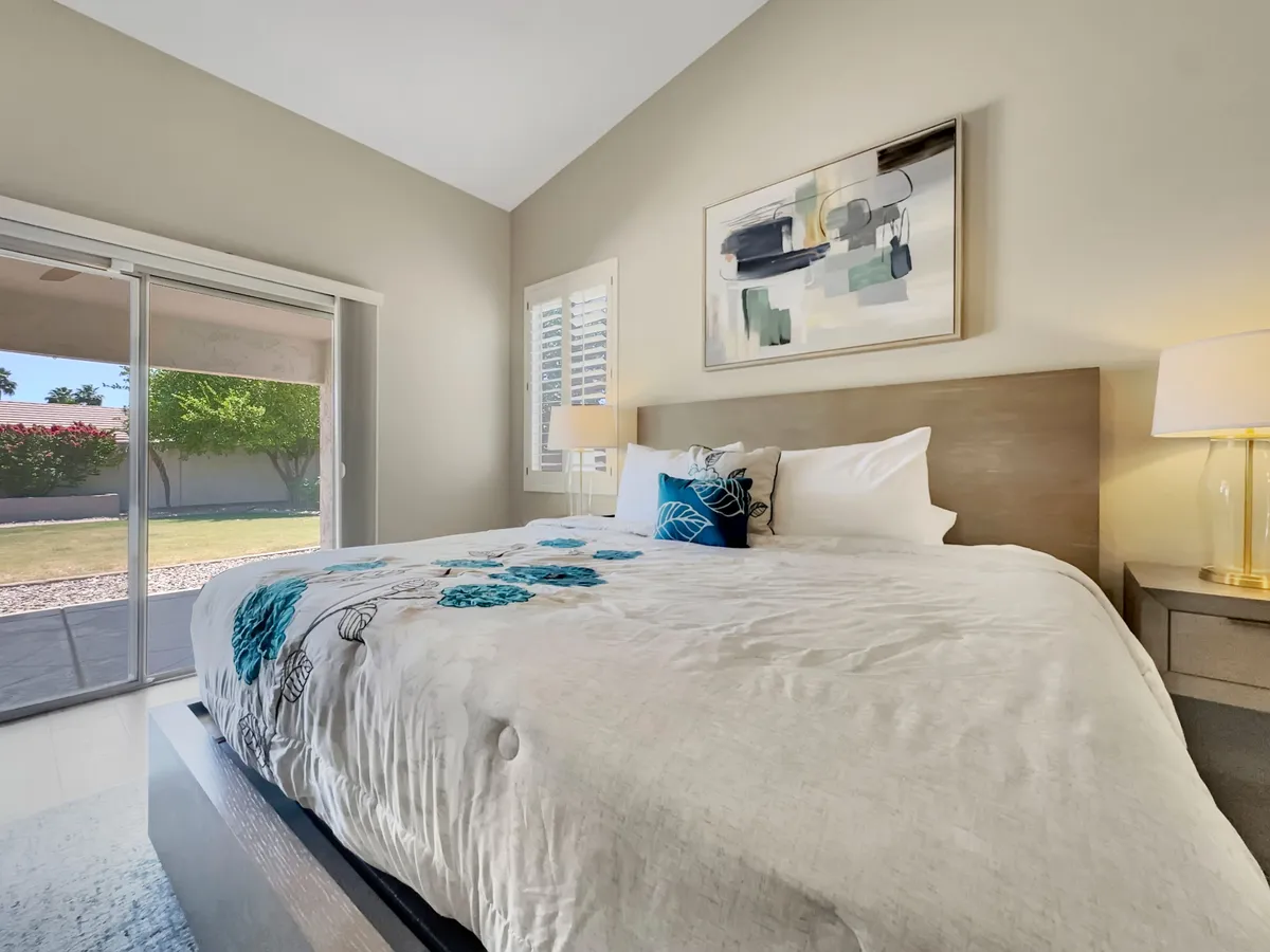 This interior image features a cozy bedroom with a bed adorned with decorative pillows, stylish artwork, and bright natural light streaming through a large sliding glass door that leads to an outdoor area. The design feels peaceful and connects indoor living to the outside.