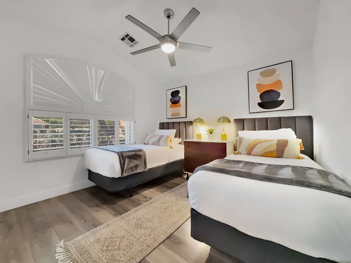 A twin/flexible bedroom with two modern beds, each adorned with fresh linens and bedside lamps. A window with shutters allows for privacy and gentle daylight, while wall art and nightstands add a welcoming touch. The space offers adaptability for guests and families.
