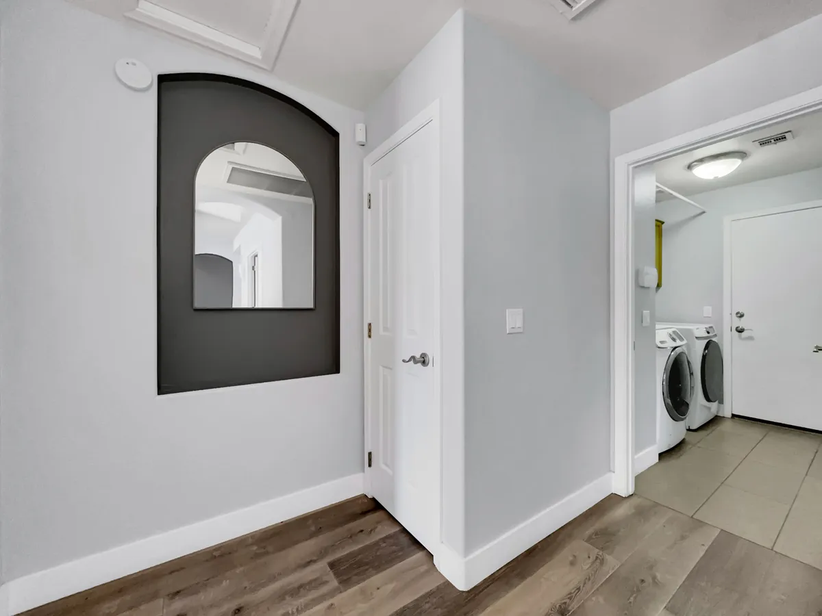 This image shows a laundry area featuring a washer and dryer with a door leading outside. The space is bright, with a decorative arched mirror and functional accents that suggest easy access between the interior and outdoors.
