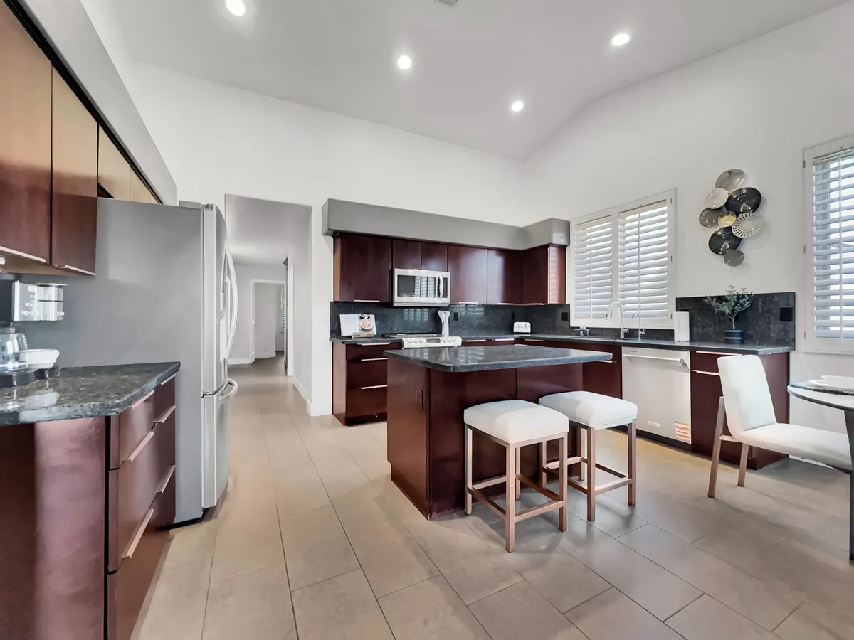 A modern kitchen featuring a prominent island, stainless steel appliances, granite countertops, and ample natural light from a window with plantation shutters. The scene suggests an inviting space perfect for both cooking and socializing.