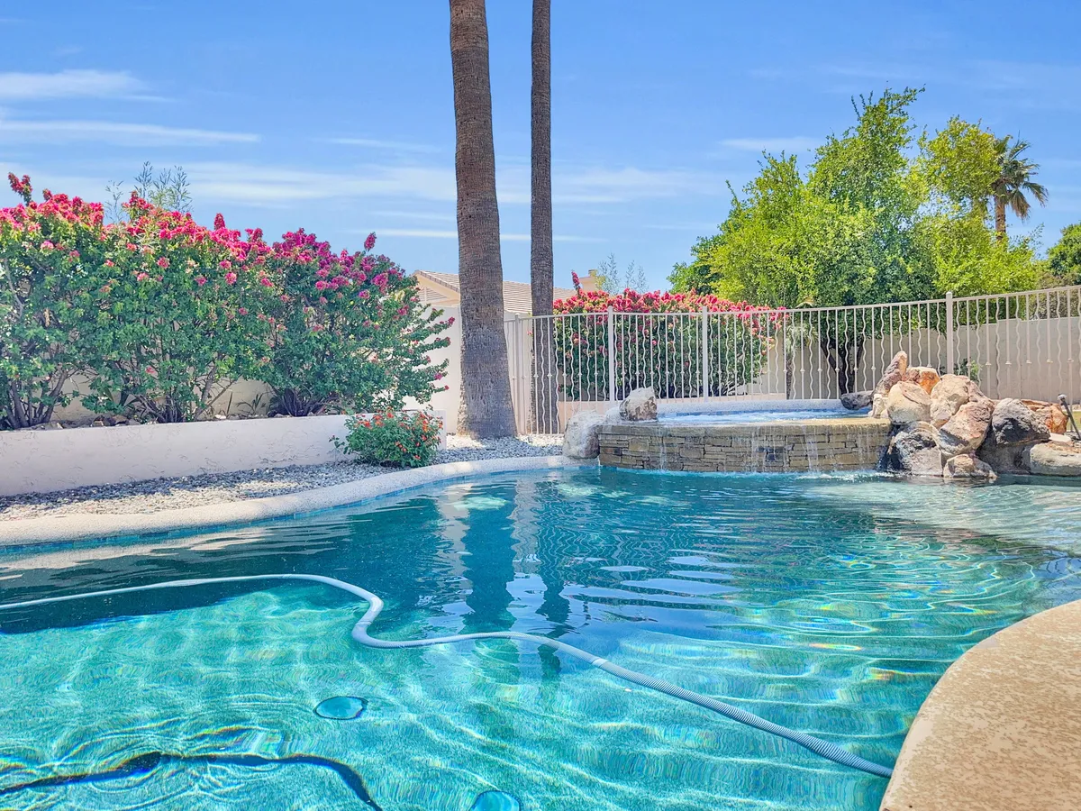 This image showcases a backyard view featuring the swimming pool set amid palm trees, beautifully landscaped flower beds, and a dedicated spa area. The calming blue of the water is contrasted by the vibrant greenery and floral accents, creating a resort-like atmosphere. The spa is positioned at poolside, and the overall layout offers plenty of open space for relaxation or entertaining.