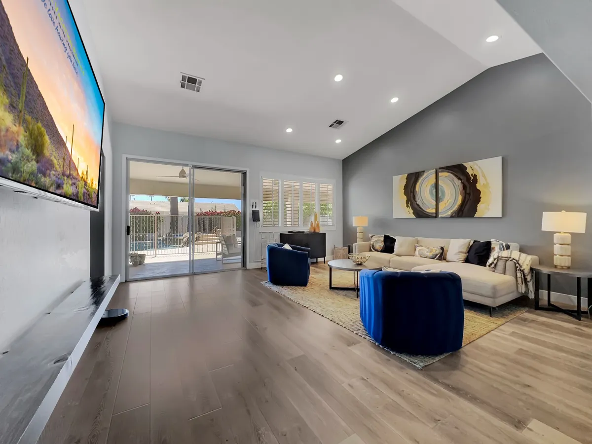 This image showcases a contemporary living room with a large television displaying a scenic image, a comfortable sofa adorned with decorative pillows, and blue accent chairs. Artwork on the wall and a sliding glass door leading to an outdoor area provide both style and a flood of natural light, echoing the open, sunlit ambiance described in the section.