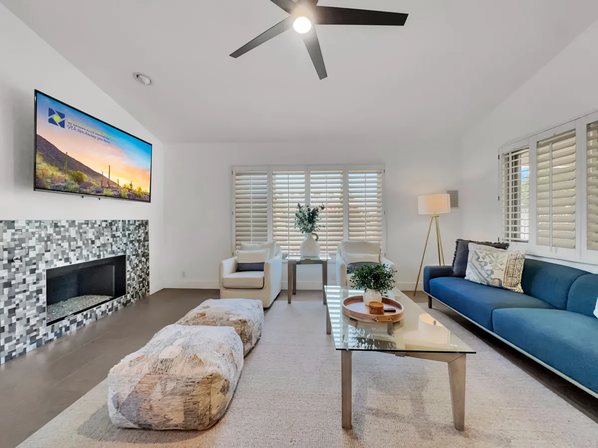 The image features a modern, cozy living room with a stylish sofa, coffee table, fireplace, television, and a touch of greenery. Natural light fills the space, and the seating is arranged for both comfortable conversation and relaxation in front of the fire or TV.
