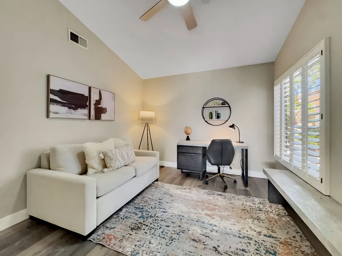 A bright, calm living space featuring a sofa, a dedicated desk with a lamp, stylish rug, and artwork. The setup suggests a comfortable place for both relaxation and productivity, with the desk area ideal for remote work or planning business outings.
