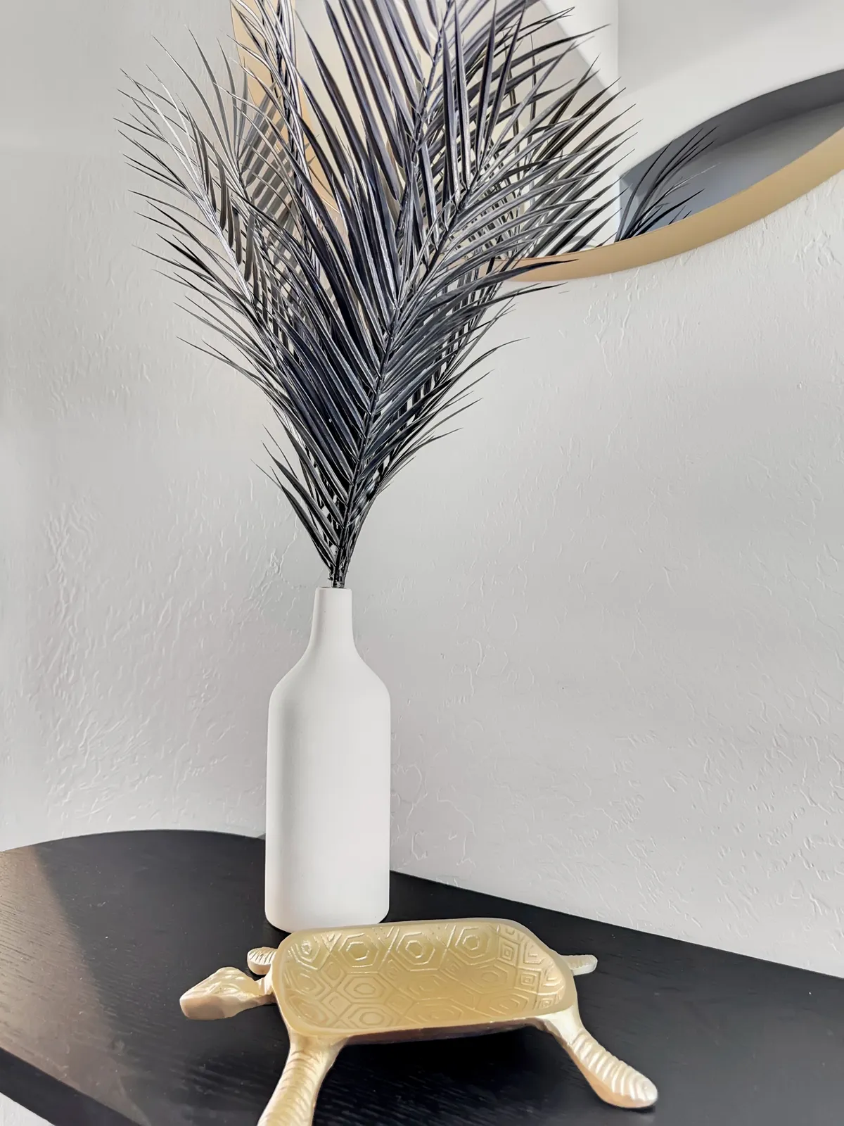 A view of the entryway with a tall black palm leaf arrangement in a white vase and a gold turtle decorative piece atop a stylish console. The decor sets a welcoming tone, inviting guests into the heart of the home.